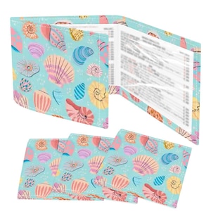 May include: A wallet and coasters set with a seashell pattern. The wallet is open, revealing pages. The coasters are arranged below. The design features various seashells in pink, yellow, and blue on a light blue background.