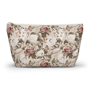 Puede incluir: Un estuche con cremallera que presenta un fondo beige con un patrón repetitivo de flores blancas y rojo oscuro, aves de cuello largo y mariposas. El diseño incluye elementos botánicos detallados y figuras de aves en toda la superficie.