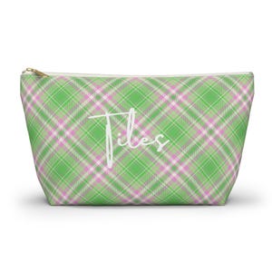 Puede incluir: Un estuche con cremallera que presenta un patrón de cuadros en verde, blanco y rosa. La palabra "Tiles" está impresa en letra cursiva blanca en el centro del estuche. La bolsa tiene un tirador de cremallera de metal dorado en el borde superior.