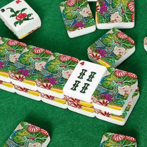 May include: A collection of white and colorful Mahjong tiles with a green felt background. The tiles feature floral and dragon designs in green, pink, and blue. Some tiles display the number four and the character "II".