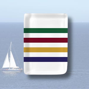 May include: A rectangular white ceramic tray featuring four horizontal stripes in dark green, deep red, golden yellow, and navy blue. The tray has rounded corners and a smooth, glossy finish, presenting a clean and simple design for holding small items.