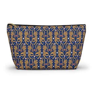 Peut inclure: Une pochette zippée bleu marine avec un motif répété de guépards dorés, de fleurs roses et d'étoiles blanches. La pochette a une fermeture éclair dorée et une forme rectangulaire.