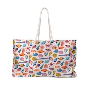 May include: A large tote bag with a white rope handle. The bag features a repeating pattern of colourful seashells, starfish, and other marine life on a light pink background. The bag is rectangular and appears to be made of fabric.