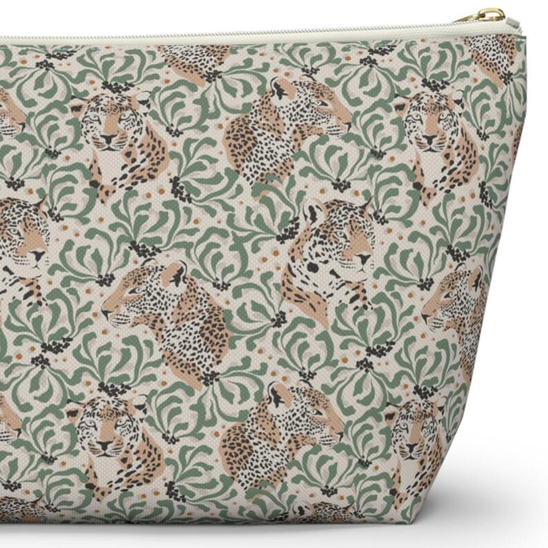 May include: A beige zippered pouch featuring a repeating pattern of stylized jaguar faces and green floral elements. The jaguar's fur is tan with black spots. The pouch has a gold-colored zipper.