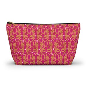 Peut inclure: Une trousse à fermeture éclair rose avec un motif répété de guépards dorés et d'éclats d'étoiles blancs. La trousse a une fermeture éclair noire et est conçue pour les cosmétiques ou les voyages.
