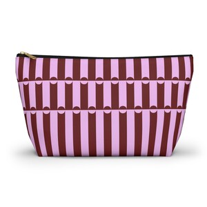 Puede incluir: Un neceser rectangular con un patrón repetitivo de rayas verticales granates y rosa claro. La bolsa tiene una cremallera negra y un tirador dorado. La bolsa está diseñada para guardar maquillaje y otros productos de belleza.