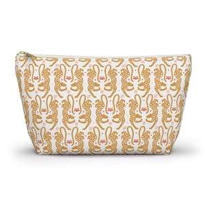 Peut inclure: Une trousse de maquillage blanche avec un motif répété de guépards dorés et d'accents floraux roses. Le sac a une fermeture éclair dorée et une forme rectangulaire, idéale pour ranger le maquillage ou les articles de toilette.