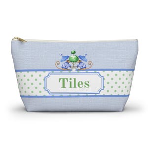 May include: A light blue zippered pouch with a textured fabric finish. The design features a central white rectangular label with the text Tiles in green, surrounded by a green polka dot band and two monkeys flanking a potted topiary tree.