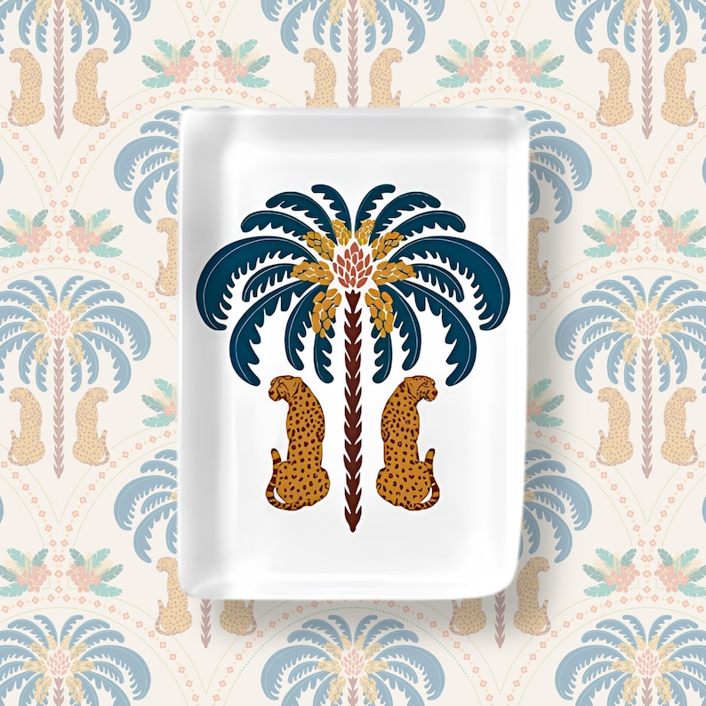 May include: A rectangular white ceramic trinket tray featuring a central graphic illustration of a dark blue palm tree with golden fruit and two spotted leopards sitting on either side of the brown trunk, rendered in a flat, symmetrical design.