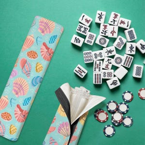 May include: A turquoise Mahjong set with a seashell-patterned case, scattered tiles, and poker chips on a green surface. The tiles are white with black and red symbols. The case has a zipper and a black trim.
