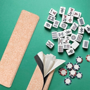 Puede incluir: Un juego de Mahjong completo con fichas blancas con caracteres y símbolos chinos, dispuestas sobre una superficie verde. El juego incluye un estuche beige con estampado floral y fichas de colores. El estuche está abierto, revelando el interior.