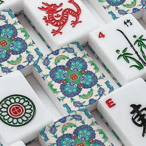 May include: Close-up of a Mahjong set with white tiles. Some tiles feature red dragon, green bamboo, and black Chinese characters. Other tiles have a floral pattern in blue, green, and white.