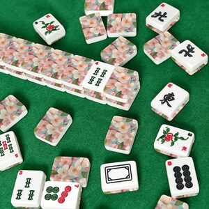 May include: A collection of white Mahjong tiles with various symbols and floral patterns, scattered on a green surface. Some tiles feature pink and white flower designs, while others display Chinese characters and dots.