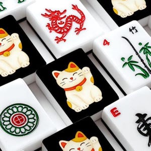 May include: Close-up of a set of Mahjong tiles. The tiles feature various designs, including a red dragon, a lucky cat, and bamboo. The tiles are white with black accents and colorful illustrations.