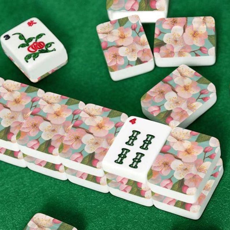 May include: A collection of white Mahjong tiles with floral designs. The tiles feature pink cherry blossoms on a light blue background. Some tiles display Chinese characters and numbers. The game pieces are arranged on a green surface.