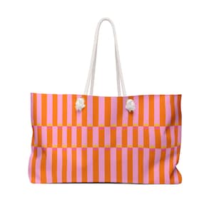 May include: A large tote bag with a vibrant, colorful design. The bag features vertical stripes in shades of pink and orange, with small yellow squares. It has white rope handles and white decorative knots. This bag is perfect for carrying beach essentials or everyday items.