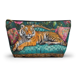 May include: A zipped pouch with a striking design of a tiger lounging on a teal, button-backed sofa, adorned with floral cushions. The background showcases tropical foliage and pink blooms, offering a luxurious feel.