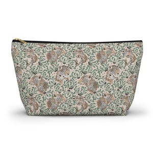 Peut inclure: Une pochette beige zippée avec un motif répété de visages de léopards et d'éléments floraux verts. La pochette a une fermeture éclair noire et une tirette dorée. Le design général évoque une esthétique vintage.