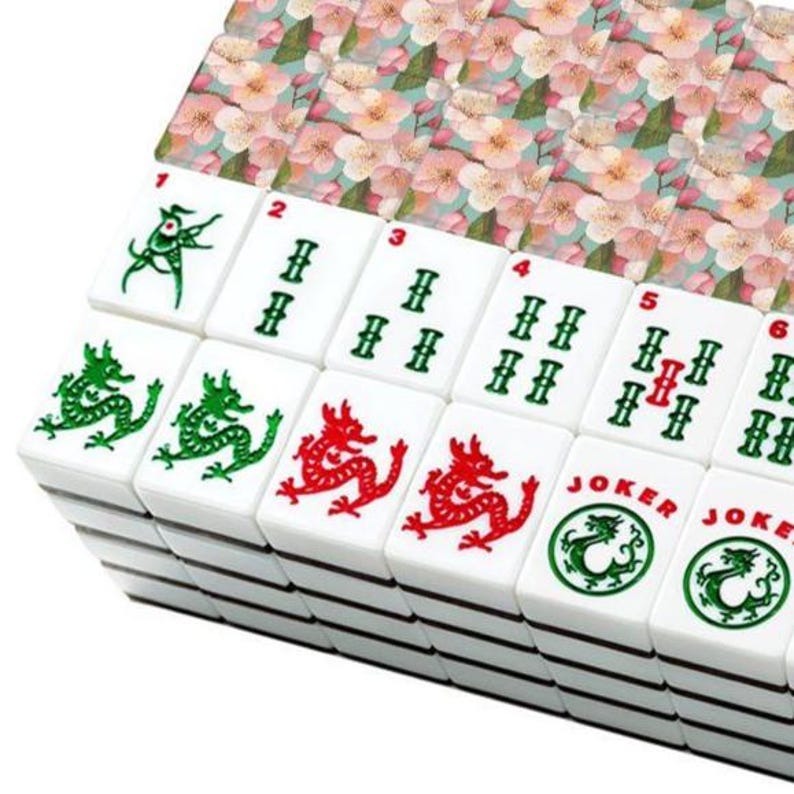 May include: A set of white Mahjong tiles with green, red, and black symbols and numbers. The tiles are stacked, with some showing dragon and joker designs. The background features floral patterned cards.