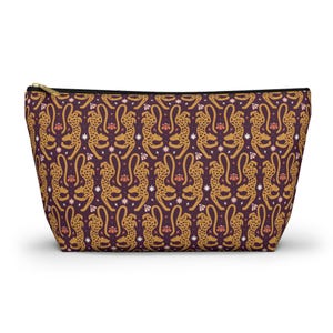 Peut inclure: Une pochette zippée avec un motif répété de guépards dorés, de fleurs et d'étoiles sur un fond violet foncé. La pochette a une fermeture éclair noire et est conçue pour tenir debout.