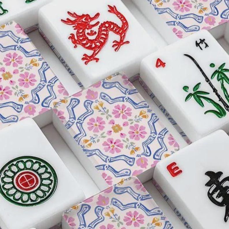 May include: A collection of rectangular mahjong tiles featuring white surfaces with various designs. Some tiles display a red dragon, a green bamboo stalk, a circular green and red pattern, or black Chinese characters. Others have a floral pattern.
