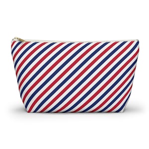 May include: A trapezoidal zippered pouch featuring a repeating diagonal striped pattern in red, white, and navy blue. The pouch has a gold-toned metal zipper pull at the top edge, and the overall shape is wider at the base than at the top opening.