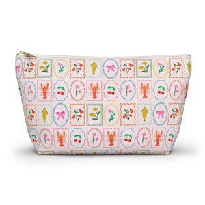 May include: A zipped pouch featuring a repeating pattern of framed illustrations. The illustrations include a lobster, cherries, a fish, a bow, and flowers. The pouch has a light pink background and a gold zip.