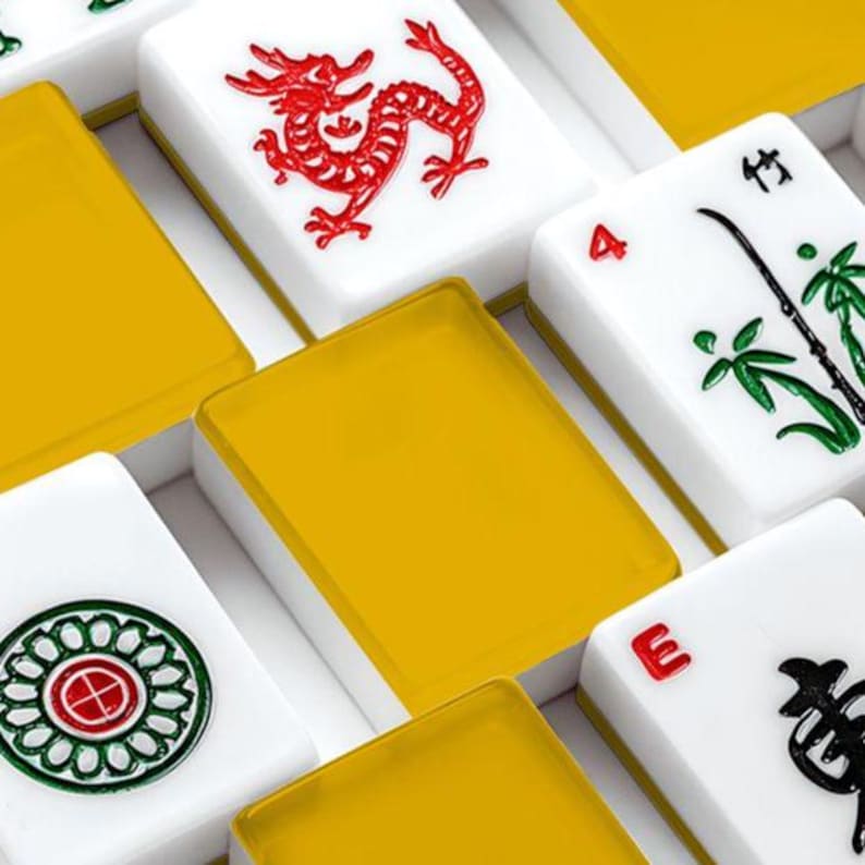 May include: Close-up of a Mahjong set with white tiles and yellow accents. The tiles feature red dragon, green bamboo, and other symbols. The set is arranged in a box, showcasing the game's components.