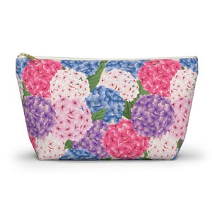 Puede incluir: Un estuche trapezoidal con cremallera que presenta un estampado integral de racimos de flores de hortensia en tonos rosa, morado, azul y blanco, intercalados con hojas verdes sobre un fondo claro, con una cremallera de metal dorado.