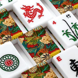 Puede incluir: Una colecci&oacute;n de fichas de mahjong rectangulares con superficies blancas y dise&ntilde;os grabados. Algunas fichas muestran dragones rojos, tallos de bamb&uacute; verdes o patrones circulares, mientras que otras presentan un estampado floral y de drag&oacute;n multicolor.