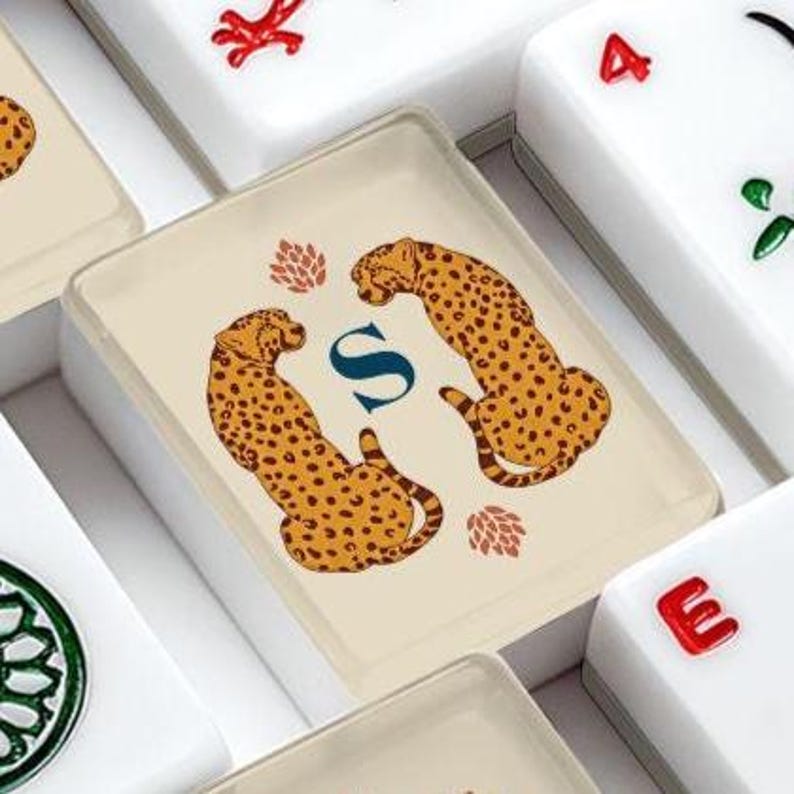 May include: A rectangular mahjong tile featuring a cream-colored face with a central dark blue letter S flanked by two illustrated cheetahs in profile. The cheetahs are yellow with black spots, and two small reddish-brown floral motifs are included.