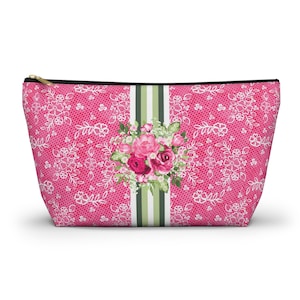 May include: A pink zippered cosmetic bag with a white lace pattern. The bag features a central vertical stripe with green and white stripes, and a floral bouquet of pink and red roses.