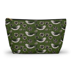 Peut inclure: Une pochette zippée avec un motif répété d'oiseaux blancs et noirs stylisés, d'éclats d'étoiles et de formes rayées sur un fond vert foncé. La pochette a une fermeture éclair dorée et une forme rectangulaire.