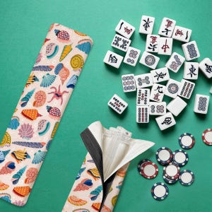 Puede incluir: Un juego de Mahjong con fichas, fichas y una funda estampada sobre una superficie verde. La funda presenta un diseño de conchas marinas y criaturas marinas. Las fichas blancas tienen símbolos negros y rojos. Las fichas tienen bordes rojos, blancos y azules.