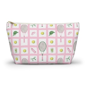 Könnte beinhalten: Eine Reißverschlusstasche mit einem rosa-weiß karierten Muster. Das Muster umfasst Tennisschläger, Tennisbälle, Visiere, Wasserflaschen und Getränke. Die Tasche hat einen goldfarbenen Reißverschluss.