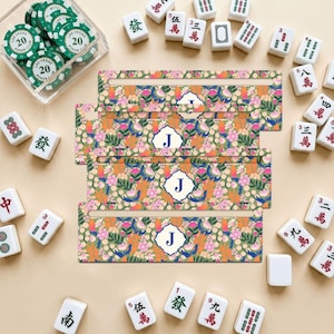 Puede incluir: Cuatro soportes rectangulares para fichas de mahjong con un colorido patrón floral de motivos botánicos rosas, naranjas y verdes. Cada soporte muestra un emblema festoneado blanco central con la letra J mayúscula en azul oscuro.