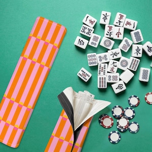 Puede incluir: Vista superior de un juego de Mahjong sobre una superficie verde. El juego incluye fichas blancas con símbolos negros y rojos, fichas de colores y una funda a rayas rosas y naranjas. La funda está abierta, revelando un interior blanco.