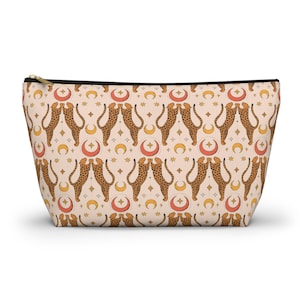 Peut inclure: Une trousse de maquillage beige avec un motif répété de guépards, de lunes, d'étoiles et de soleils. Le sac a une fermeture éclair noire et une tirette dorée. Le motif est dans les tons de marron, d'orange et de jaune.