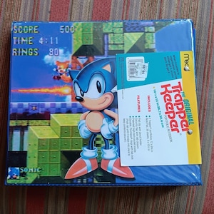 Puede incluir: Un cuaderno portafolio azul Trapper Keeper con Sonic the Hedgehog. El cuaderno mide 26,7 x 30,4 cm. La portada muestra un gráfico colorido de Sonic en una escena de videojuego, con el texto "SCORE 500, TIME 4:11, RINGS 80".