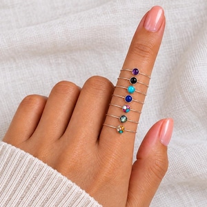 Op de afbeelding: Een set van zeven dunne metalen ringen gestapeld op een vinger, elk met een kleine ronde edelsteen. De stenen zijn paars, zwart, turquoise, blauw, veelkleurig, donkergroen en beige gespikkeld, allemaal geplaatst op een eenvoudige ringband.