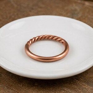 Puede incluir: Un anillo de color cobre que presenta una banda lisa en la mitad inferior y una textura trenzada similar a una cuerda en la mitad superior. El anillo es circular y tiene un acabado metálico con un tono cálido de color marrón rojizo.