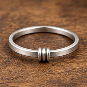 Puede incluir: Un anillo de metal plateado con un acabado mate cepillado que presenta tres pequeños anillos plateados, lisos y pulidos, colocados juntos como una sola unidad en la banda principal del anillo, creando un punto focal distintivo.