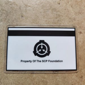 May include: A rectangular card with a clear plastic border. The card has a white background with a black stripe at the top. The card features a black logo and the text "Property Of The SCP Foundation."