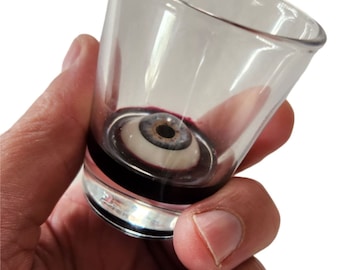 May include: A clear glass shot glass containing a fake eyeball. The eyeball is white with a blue iris and a black pupil. The shot glass is filled with a dark red liquid.