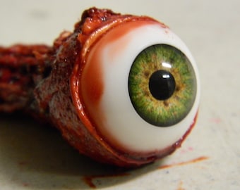 May include: A realistic fake eyeball with a green iris and a red, bloody socket.