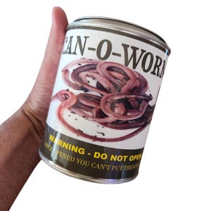 Può includere: Una lattina novità etichettata "CAN-O-WORMS" con una grafica di lombrichi. La lattina ha un'etichetta bianca con testo nero che recita "WARNING - DO NOT OPEN ONCE OPENED YOU CAN'T PUT THEM". La lattina è tenuta in mano.