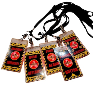 Peut inclure: Plusieurs badges d'identification en plastique transparent avec des cordons noirs. Chaque badge présente un fond noir avec des rayures de danger jaunes et le mot "CAUTION". Les badges affichent différents textes, dont "EMERGENCY", "SELF DESTRUCT", "FIRE ALARM", "PANIC BUTTON" et "RADIATION DETECTION".