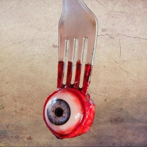 May include: A silver metal fork with four tines piercing a realistic, spherical human-like eyeball. The eyeball features a blue iris, a black pupil, and is surrounded by a textured, deep red, fleshy base that coats the lower part of the fork tines.
