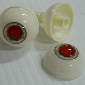 以下が含まれることがあります： 血のような赤い虹彩と濃い茶色の外側のリングが付いた、オフホワイトのプラスチック製の眼球が3つ。1つの眼球は分解されており、中空の背面と別のレンズが見えます。白い表面に置かれたアイテムです。