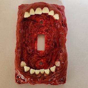 Può includere: Una placca per interruttore della luce con un design rosso, carnoso e testurizzato. La placca ha una fila di denti bianchi in alto e in basso, che ricordano una bocca.